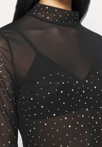 Svart genomskinlig topp med hög krage, med ett prickigt mönster av iriserande rhinestones på framsidan och en lagerdesign.