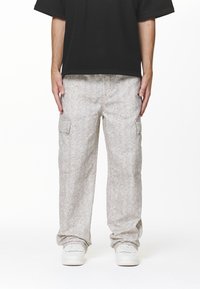Persoon in lichtgrijze geprinte cargobroek, witte sneakers en een zwart T-shirt met korte mouwen, staand met armen ontspannen langs het lichaam.