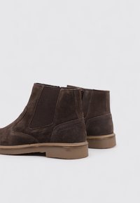 Botas Chelsea de ante marrón con paneles laterales elásticos, detalles de costura y una suela de goma baja, con una parte superior texturizada y punta redonda.