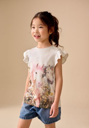 Młoda dziewczyna stojąca w jeansowych szortach i białej bluzce z falbankami na rękawach z kolorowym nadrukiem koni i kwiatów, uśmiechająca się, beżowe tło.