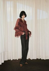 Rode blouse met paisleypatroon en ruches aan de mouwen, gecombineerd met donker wijde broek en bruine puntige schoenen; minimalistische achtergrond.