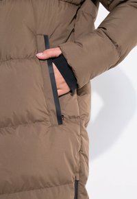 Veste matelassée marron avec un design rembourré, poche latérale avec finition noire et poignets noirs. Texture lisse et lignes épurées.