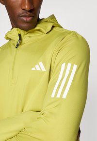 Hellgrüner Sporthoodie mit einer halben Reißverschluss-Kragen, strukturiertem Stoff und drei weißen Streifen am linken Ärmel. Logo auf der Brust.