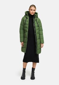 Woman wearing a mid-length green puffer coat over a black ribbed knit dress and black ankle boots, standing against a plain background.