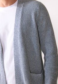 Grå teksturert cardigan laget av strikkemateriale, med åpen front, lange ermer og en enkelt lappelomme på venstre side.