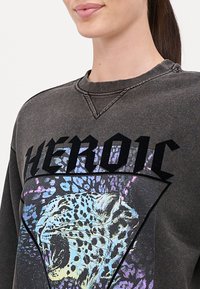 Tummanharmaa huppari, jossa on suuri graafinen kuvasto karjuvasta leopardista ja rohkeasta "HEROIC"-tekstistä mustana, kuluneen ja tekstuurilla varustetun viimeistelyn kera.