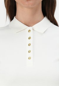 Bílá polo košile s límečkem a šesti zlatými knoflíky vepředu. Materiál se zdá být texturovaný a lehký.