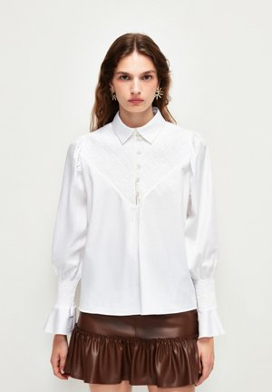 Camicia bianca con colletto strutturato, chiusura con bottoni e pannellatura testurizzata. Presenta maniche a sbuffo con polsini arricciati, indossata con una gonna marrone.