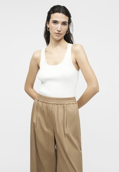 Νεαρή γυναίκα με σκούρα μαλλιά φοράει σκουλαρίκια κρίκους, λευκό αμάνικο τοπ και μπεζ παντελόνι φαρδιάς γραμμής, στέκεται με τα χέρια πίσω από την πλάτη.