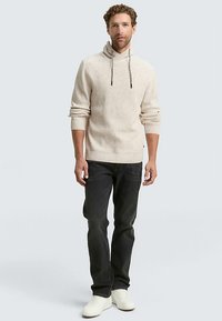 Beige stickad hoodie med trattkrage och dragsko, kombinerad med mörka jeans och vita sneakers. Har subtil textur och en avslappnad passform.