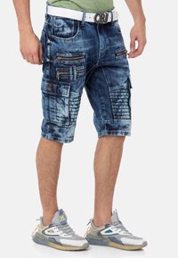 Blaue Denim Cargo-Shorts mit mehreren Reißverschlusstaschen, kontrastierenden Nähten und einem strukturierten Design. Kombiniert mit grauen Sneakers.