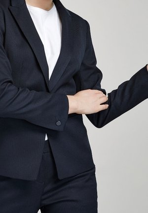 Blazer bleu marine foncé en tissu texturé, avec une fermeture à un bouton et des revers en pointe. Chemise blanche en dessous. Bras légèrement ployé.