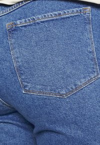 Džínové kalhoty ukazující detail na zadní kapsu. Vlastnosti zahrnují modrou látku, oranžové prošívání a mírně vybledlý vzhled.