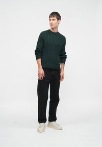 Σκούρο πράσινο πλεκτό πουλόβερ με υφή καλωδίου, στρογγυλή λαιμόκοψη και ριμπ μανσέτες, συνδυασμένο με μαύρο παντελόνι και λευκά αθλητικά παπούτσια.