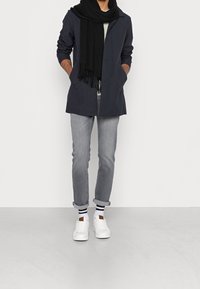 Marinblå kappa, grå jeans, vita sneakers och en svart scarf. Outfitten har en modern, minimalistisk design med uppvikta ärmar och randiga strumpor.
