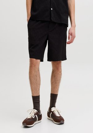 Homme portant un short noir, des chaussettes foncées et des baskets marron, debout devant un fond clair uni.