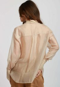 Blusa traslucida color pesco chiaro con maniche lunghe, colletto con bottoni e vestibilità morbida. Famosa per la sua morbidezza e trasparenza.