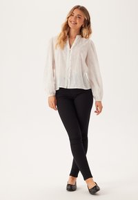 Witte blouse met lange mouwen, opgedrukte oogpatronen, knopen aan de voorkant en een ontspannen pasvorm. Gecombineerd met zwarte slim-fit broek en zwarte platte schoenen.