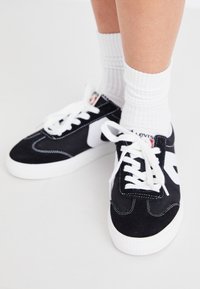 Czarne zamszowe sneakersy z białymi akcentami, z okrągłym noskiem, grubymi białymi sznurowadłami i teksturowaną gumową podeszwą. Noszone z białymi skarpetkami do kostek.
