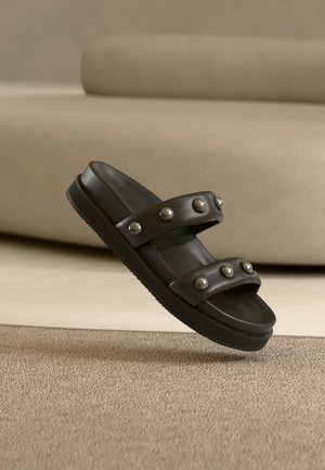 Juoda slidžių tipo sandalė su dviem paminkštintomis juostelėmis, kiekviena papuošta šešiais sidabriniais apvaliais kniedėmis, padėta ant kreminės kilimo su atitinkančiu sofa fone.