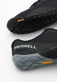 Scarpe sportive Merrell in nero e grigio con suole vibram testurizzate, evidenziate dai loghi del marchio sul tallone e sulla suola.