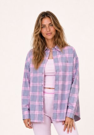 Femme aux cheveux longs et ondulés portant une chemise à carreaux bleue et rose boutonnée sur un crop top rose clair et un pantalon taille haute assorti.