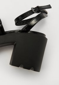 Black heeled sandal featuring a glossy patent leather upper, adjustable ankle strap, and a chunky block heel with a matte finish.