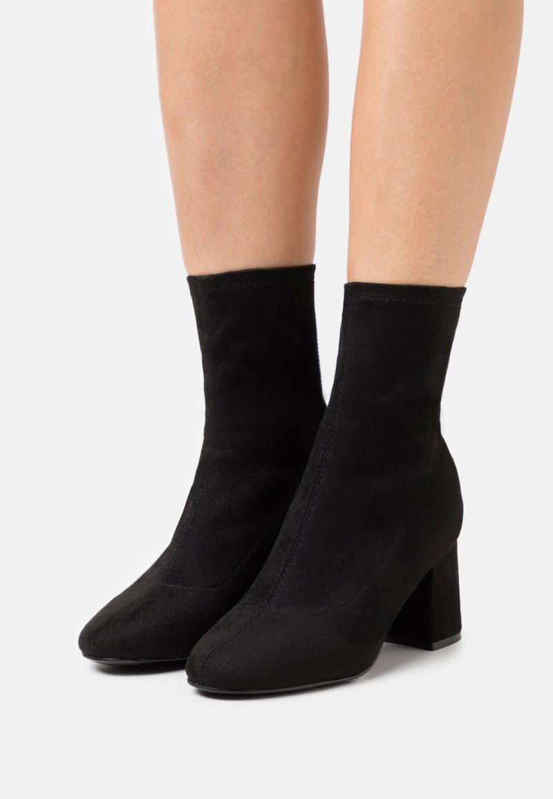 Paire de bottines en suède noir à talons carrés portées sur des jambes nues sur un fond blanc uni.
