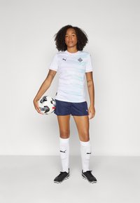 White athletic jersey with a wave pattern, navy shorts, white socks, and black cleats. Holding a black and white soccer ball.
