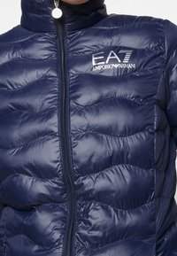 Navy blue puffer jacket with quilted pattern, high collar, front zipper, and EA7 Emporio Armani logo on the left chest. Smooth texture.