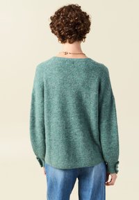 Ulltröja i teal, med avslappnad passform, ribbad textur, sänkta axlar och knappdetaljer på ärmarna. Matchad med blå jeans.