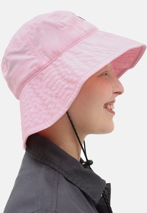 Personne souriante portant un chapeau seau à large bord rose avec une sangle réglable sous le menton et une veste gris foncé, vue de profil droit.