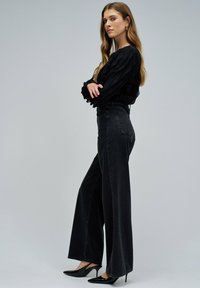 Vaqueros negros de pierna ancha con cintura alta, combinados con una blusa negra texturizada con mangas acampanadas. Modelo que lleva zapatos de tacón negros.