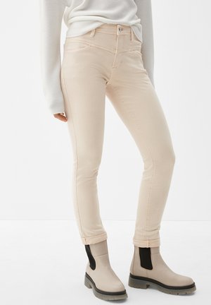 Beige slim-fit broek met omgeslagen boord, gedragen met beige en zwarte enkellaarzen en een witte geribde trui met lange mouwen.