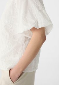 Weiße Bluse mit einem gestickten Blumenmuster, lockeren kurzen Ärmeln und einem weichen, strukturierten Stoff. Die Trägerin hat ihre Hand in einer Tasche.