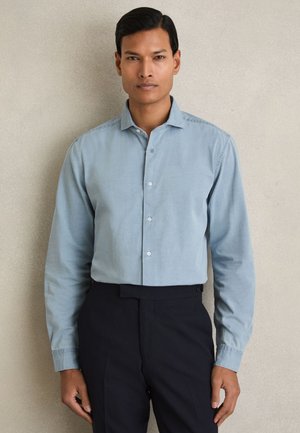 Férfi világoskék gombos ingben és fekete nadrágban, egy sima bézs fal előtt áll.