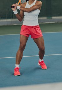 Pantalones cortos deportivos de color rosa brillante combinados con una camiseta sin mangas blanca. El sujeto sostiene una raqueta de tenis y lleva zapatillas de entrenamiento de color rosa neón.