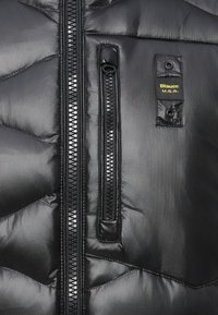 Giacca a vento nera con finitura lucida, caratterizzata da motivi a cucitura diagonale, due tasche con zip e un'etichetta gialla con la scritta "Blauer, U.S.A."
