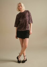 Majica s uzorkom leoparda u smešnim i crnim tonovima, slobodnog kroja s kratkim rukavima, uparena s crnom omotnom suknjom i crnim cipelama s vezicama.