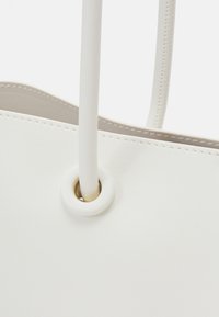 Zbliżenie na narożnik białej skórzanej torebki z przeszyciem oraz paskiem przeplecionym przez okrągły pierścień wzmacniający.