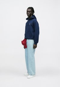 Σκούρο μπλε φούτερ με κουκούλα, με μπροστινή τσέπη και κορδόνια· ανοιχτό μπλε τζιν παντελόνι με φαρδιά πόδια· κόκκινο καπέλο κρατημένο στη μέση· λευκά αθλητικά παπούτσια.