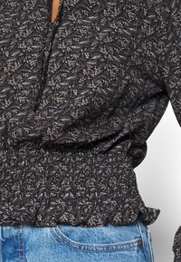 Blusa nera con motivo floreale in grigio e marrone, dotata di orlo arricciato e cintura elastica, abbinata a jeans in denim blu.