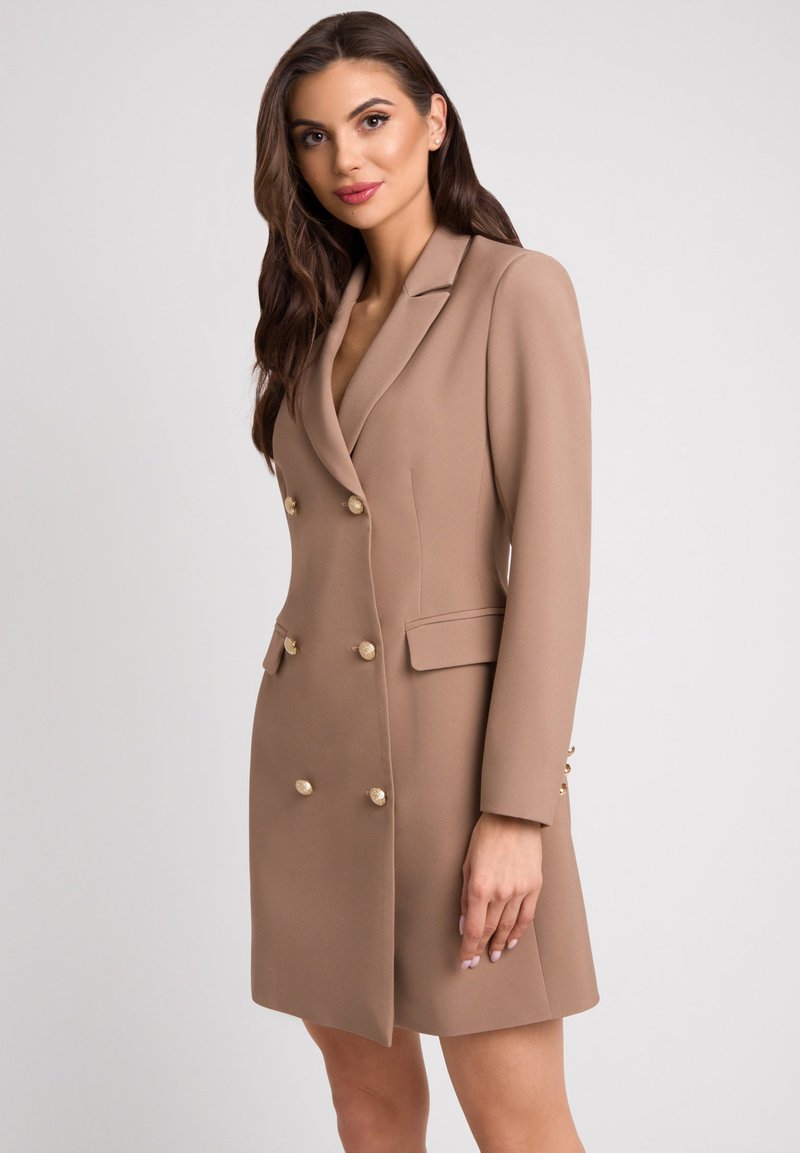 Dwurzędowa sukienka żakietowa z jasnobrązowego materiału, z złotymi guzikami, klapami i bocznymi kieszeniami, o dopasowanym kroju.