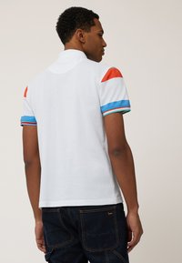 Witte polo shirt met oranje, blauwe en turquoise gestreepte schouderdetails. Korte mouwen en een kraag, gemaakt van een gladde stof.