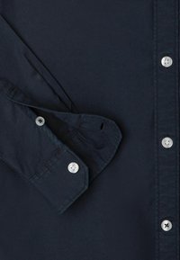 Tummansininen nappikauluspaita puuvillakankaasta, jossa on hopeanväriset napit ja rakenteellinen kaulus, pitkät hihat ja taitettavat hihansuut.