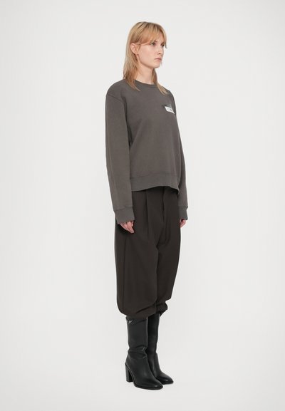 Mujer de pie de perfil, vistiendo un sudadera oscura de gran tamaño, pantalones holgados oscuros y botas de tacón negras, contra un fondo blanco liso.