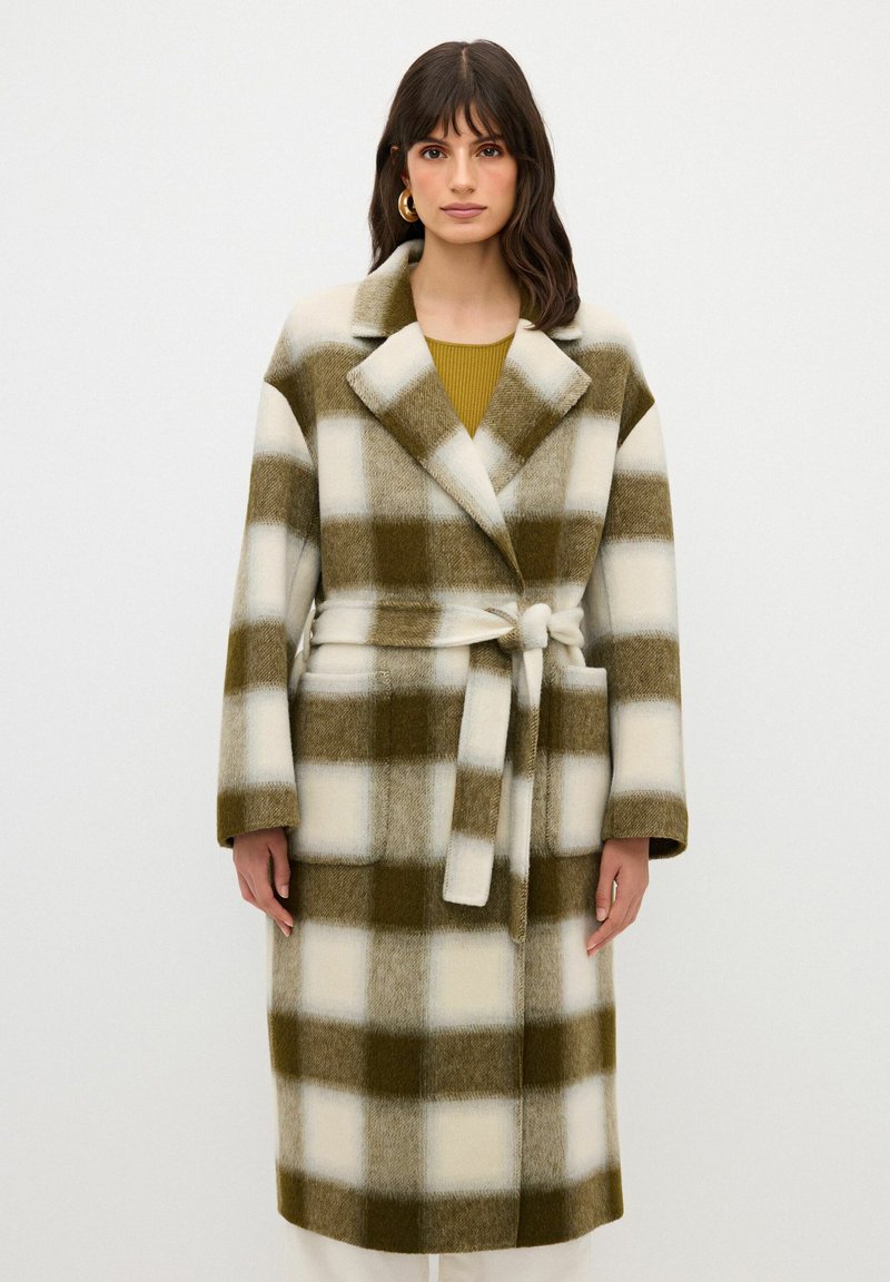 Cappotto lungo a motivi a scacchi in verde oliva e crema, realizzato in tessuto morbido. Presenta una cintura, colletto a ribalta e due tasche frontali.