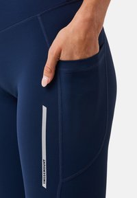 Granatowe legginsy sportowe wykonane z rozciągliwego materiału, z boczną kieszenią i odblaskowym logo. Gładka tekstura z płaskimi szwami.