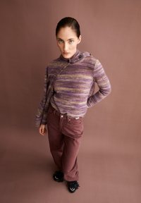 Frau mit nach hinten gekämmtem Haar, die einen lila gestreiften Pullover mit Knopfdetails, kastanienbraune Hose und schwarze Quasten-Schuhe trägt, vor einem braunen Hintergrund steht.