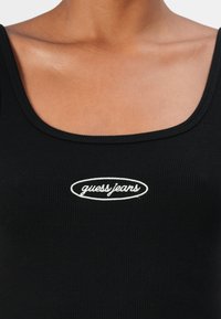 Schwarzes geripptes Tanktop mit abgerundetem Ausschnitt und einem kleinen weißen "guess jeans" Logo auf der Vorderseite.